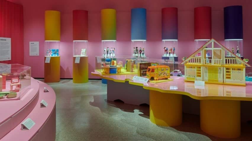 A imagem mostra a exibição da Barbie no Design Museum, destacando a importância do design do brinquedo ao longo dos anos.