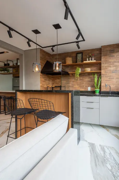 Imagem que mostra decoração de área de churrasco, incluindo pia e bancada de granito, churrasqueira de vidro e tijolos à vista.